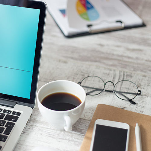 Decorative Image - "A desk with a laptop open next to a cup of coffee, glasses, smart phone and notebook and pen"