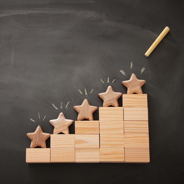 Conceptual image depicting a five-star rating system using wooden blocks and wooden stars arranged in a staircase formation on a blackboard background. Each step increases in height, symbolizing growth or progress, with chalk-drawn accents highlighting the top stars. A piece of chalk rests in the upper right corner, reinforcing the theme of evaluation and achievement.
